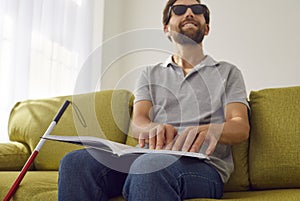 Blind smiling man sitting on sofa at home and reading tactile braille book. photo