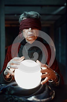 Blindfolded witch fortune teller doing predictions with illuminated crystal ball