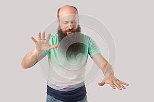 Blindness. Portrait of middle aged bald man with beard in light green t-shirt standing with closed eyes and trying to touching or