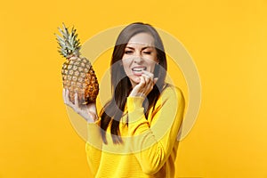 Blinking young woman in casual clothes gnawing nails, hold in hands fresh ripe pineapple fruit isolated on yellow orange