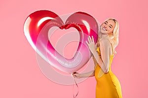 Blissful blonde woman hugging heart balloon on pink background
