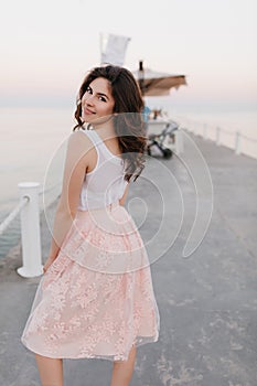 Blissful brown-haired girl in romantic attire looks over shoulder with cute smile during walk on sea pier in morning