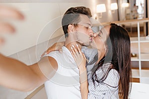 Blissful woman with elegant white manicure kissing boyfriend while he makes selfie. Handsome man with stylish haircut