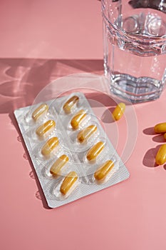 Blister with yellow pills, painkillers tablets with glass of water pink background, sun shadows