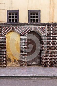 Blocked arced door, wooden door, and small windows with rusted bars on black and red bricks wall