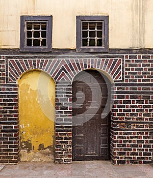 Blocked arced door, wooden door, and small windows with rusted bars on black and red bricks wall