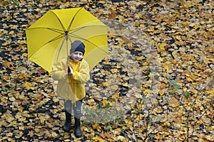 Blod boy with large yellow umbrella and raincoat on a background of yellow autumn foliage. Top view