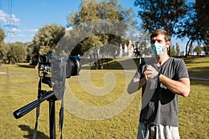 A blogger in a medical mask records a video blog on a camera in a park. Blogging concept. Vlog, remote work