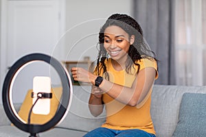 Blogging concept. Happy african american female video blogger with cellphone on ring lamp recording review on smartwatch