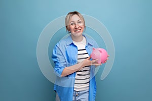 blond attractive young woman in casual attire thoughtfully holding a piggy bank with money on a blue background with