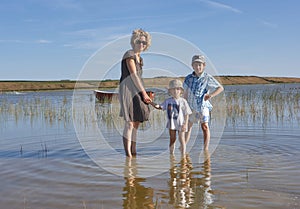 Blond beautiful mother with her two kids, standing in a lake with aquatic plants and a boat, in the background a green landscape