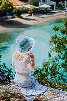 Blond beautiful woman with blue sunhat and white clothes sitting in front of blue bay of colorful tranquil village Assos