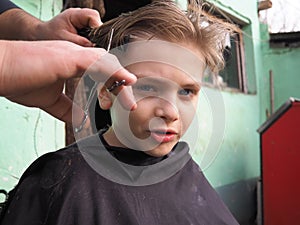 A blond boy of 8 years old gets his hair cut with scissors. Hairdresser services at home. Haircut for a child with blond hair