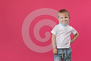 A blond boy child stands on a bright pink background, hand to side and looks mockingly and sarcastically at the camera, a mask is