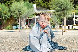 Blond boy frowns wrapped with towel sitting on beach