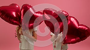 Blond boy and girl brother and sister have fun together, jumping holding red heart-shaped balloons in their hands
