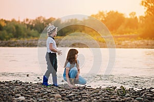 Blond boy in hat and girl playing at river shore. Kids wear rain boots. Soft focus