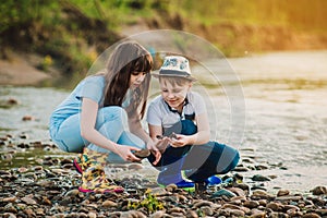 Blond boy in hat and girl playing at river shore. Kids wear rain boots. Soft focus