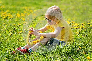 Blond boy having fun outdoor in meadow. Child sits on the grass and makes wreath of dandelions. Happy childhood