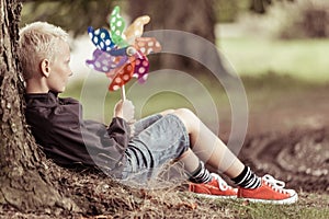 Blond boy holding colorful whirligig sits by tree