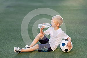 A blond boy in a sports uniform sits on a football field with a soccer ball and drinks water from a bottle, sports section.