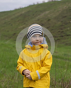 Blond boy in a yellow jacket stands on a green meadow