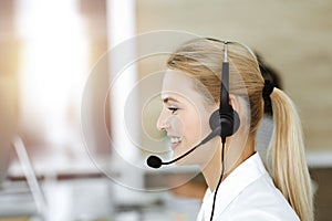 Blond business woman using headset for communication and consulting people at sunny office. Call center