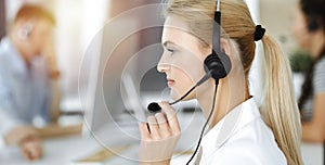 Blond business woman using headset for communication and consulting people at sunny office. Call center