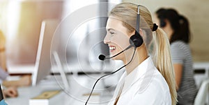 Blond business woman using headset for communication and consulting people at sunny office. Call center