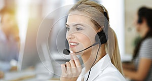 Blond business woman using headset for communication and consulting people at sunny office. Call center