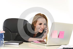 Blond businesswoman working at office laptop computer desk talking on mobile phone smiling