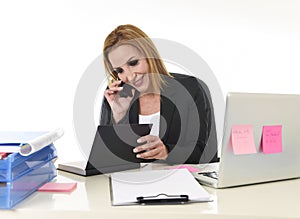Blond businesswoman working at office laptop computer desk talking on mobile phone smiling