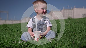 Blond caucasian boy sits on green grass in summer weather outside and quenches his thirst by drinking cold drink from