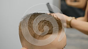 Blond caucasian boy is sitting in barber shop and hairdresser girl is cutting his hair. Child gets fashionable haircut