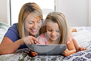Blond Caucasian mother lying on bed with her young sweet 7 years old daughter using internet on digital internet tablet pad togeth