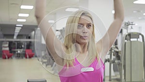 Blond Caucasian sportswoman with hazel eyes training in gym. Concentrated young woman raising hands holding dumbbells