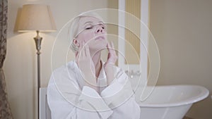 Blond Caucasian woman applying face cream. Portrait of beautiful adult lady taking care of face skin in bathroom. Beauty