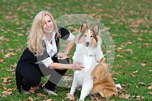 Blond Caucasian woman is sitting on a green grass and fall foliage in a park with collie dog