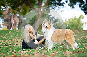 Blond Caucasian woman is sitting on a green grass and fall foliage in a park with collie dog