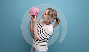 blond cheerful girl in casual attire carefully holds a piggy bank with savings on a blue background with copy space