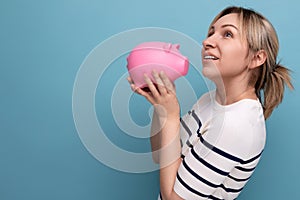 blond cheerful girl in casual attire carefully holds a piggy bank with savings on a blue background with copy space