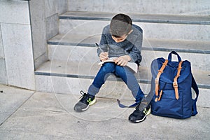 Blond child student writing on book sitting on stairs at school