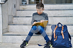 Blond child student writing on book sitting on stairs at school