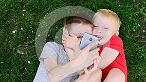 Blond and dark-haired boys lay on  grass, goofing around and taking selfies. Top view.