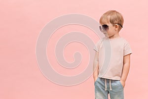 A blond European-looking child boy stands in sunglasses and looks left on a pink background