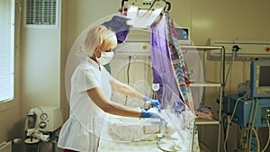 Blond female doctor in protective mask examining newborn baby in incubator. Neonatal intensive care unit.