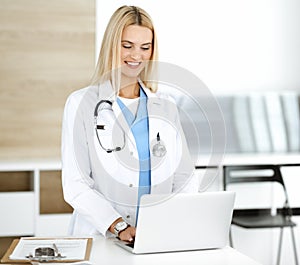 Blond female physician is lsmiling while using laptop computer. Woman-doctor at work in clinic excited and happy of her