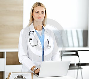 Blond female physician is lsmiling while using laptop computer. Woman-doctor at work in clinic excited and happy of her