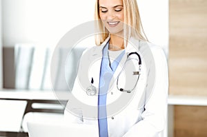 Blond female physician is lsmiling while using laptop computer. Woman-doctor at work in clinic excited and happy of her