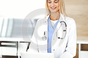 Blond female physician is lsmiling while using laptop computer. Woman-doctor at work in clinic excited and happy of her
